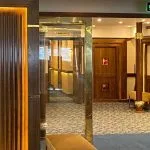 Dark wood paneled hallway entrance with bright accent lighting, gold trim, and a blue patterned carpet leading to cruise cabins.