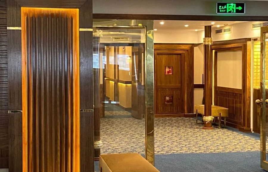 Dark wood paneled hallway entrance with bright accent lighting, gold trim, and a blue patterned carpet leading to cruise cabins.