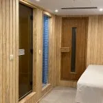 Wood-paneled spa room with a glass sauna door, a blue mosaic shower, a wooden massage table, and a sealed wooden door.