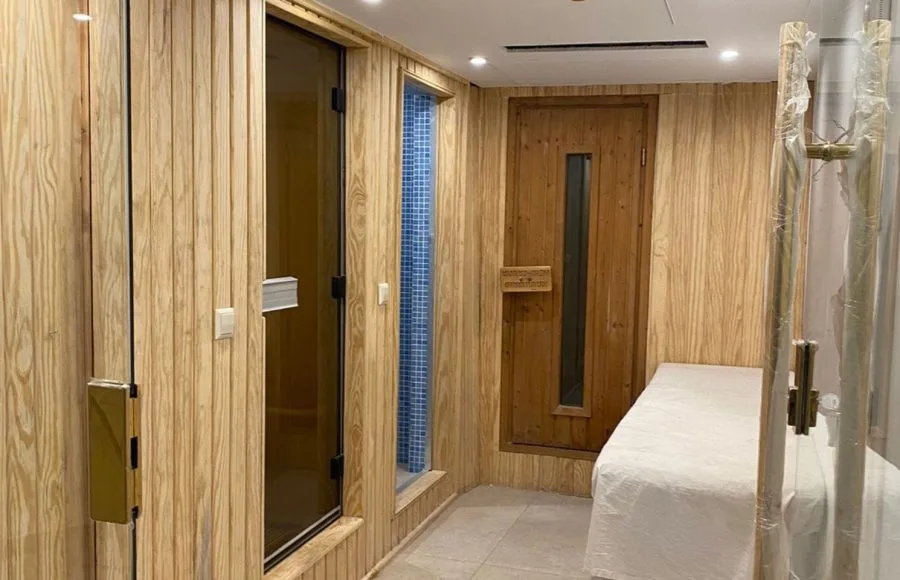Wood-paneled spa room with a glass sauna door, a blue mosaic shower, a wooden massage table, and a sealed wooden door.