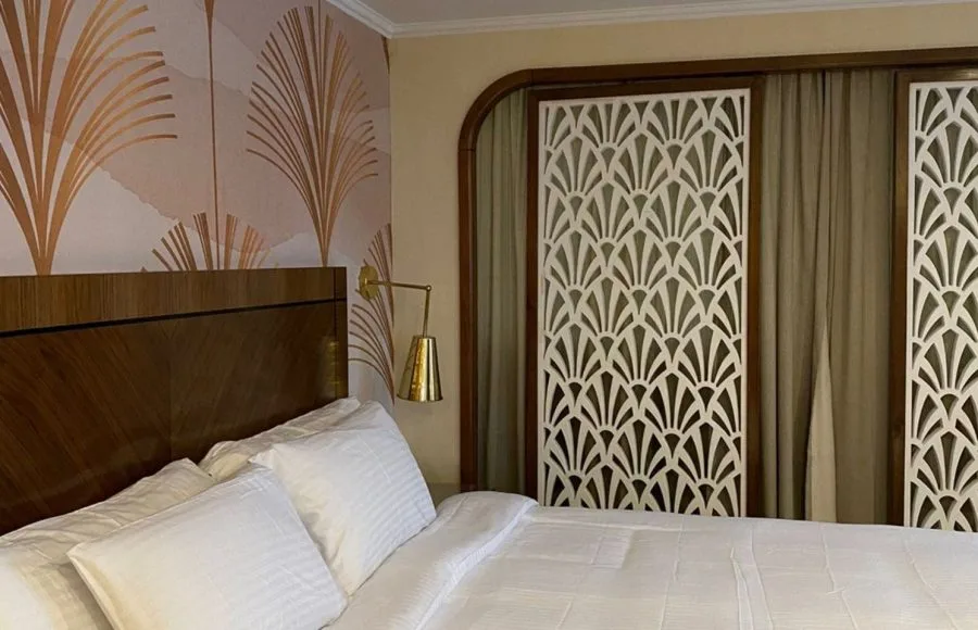 Cruise cabin wardrobe doors with white Art Deco lattice screens next to a bed with Art Deco fan pattern wallpaper.