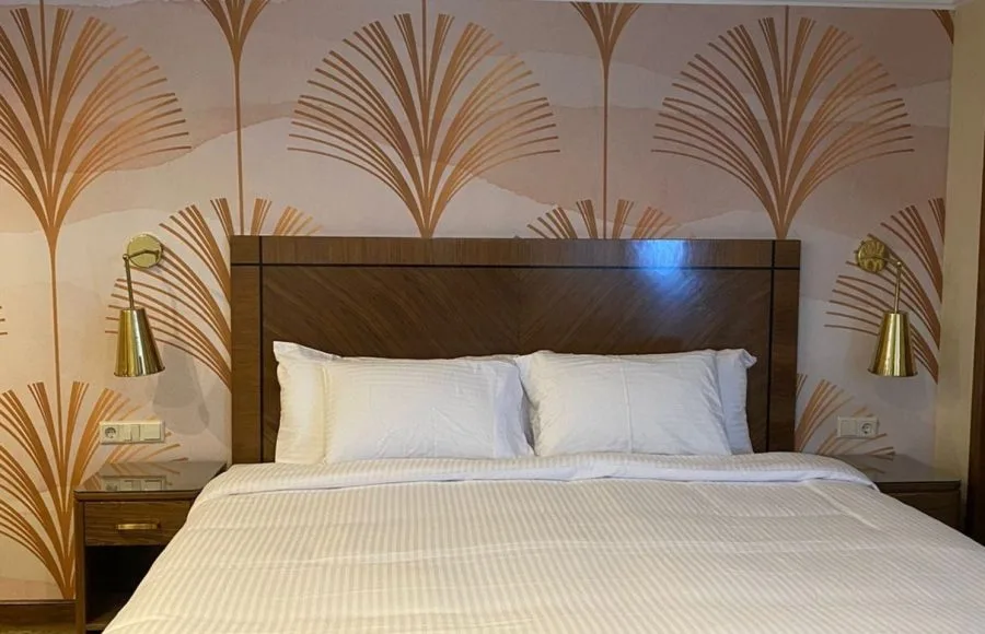 Double bed in a cruise cabin with a dark wood headboard, brass wall lamps, and Art Deco-style fan pattern wallpaper.