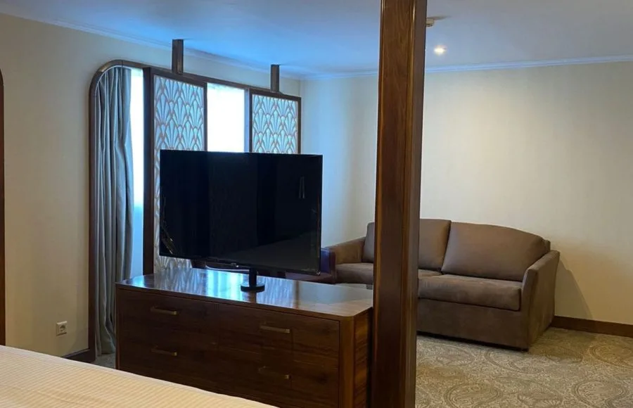 Cruise suite with a sofa sitting area separated from the bed by a dark wood TV stand/divider.