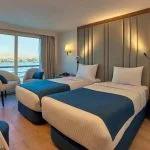 Elegant cabin with a double bed, blue accents, and floor-to-ceiling windows overlooking a river.