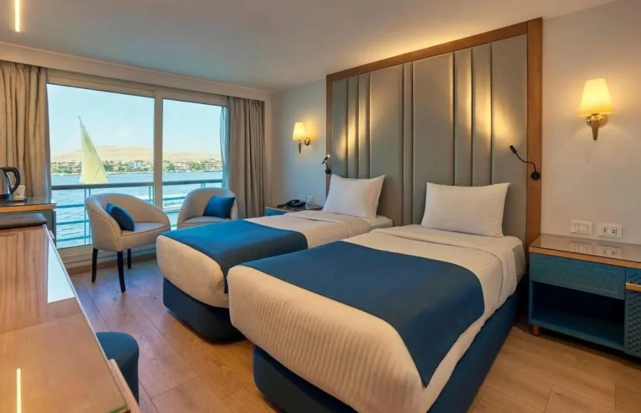 Elegant cabin with a double bed, blue accents, and floor-to-ceiling windows overlooking a river.