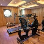 Small cruise ship gym with a treadmill, elliptical, and exercise bike, featuring wood paneling.