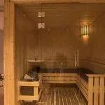 Clean, wooden sauna room with tiered benches, a glass door, and adjacent massage room visible.