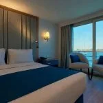 Modern, blue and white cruise cabin with a large bed, two armchairs, and floor-to-ceiling windows with a river view.