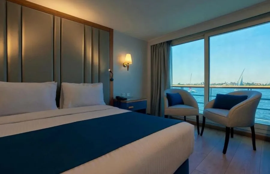 Modern, blue and white cruise cabin with a large bed, two armchairs, and floor-to-ceiling windows with a river view.