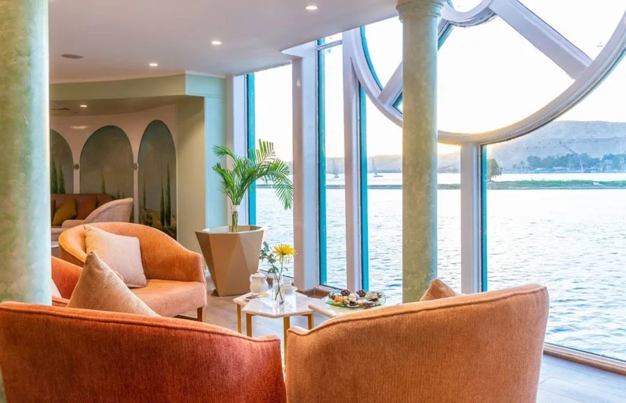 A bright and inviting lounge on the Sonesta Moon Goddess Nile Cruise ship, with comfortable seating and large windows offering a panoramic view of the Nile.