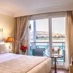A luxurious suite on the Sonesta Moon Goddess Nile Cruise ship, featuring a comfortable bed, a large window, and a private balcony overlooking the scenic Nile River.