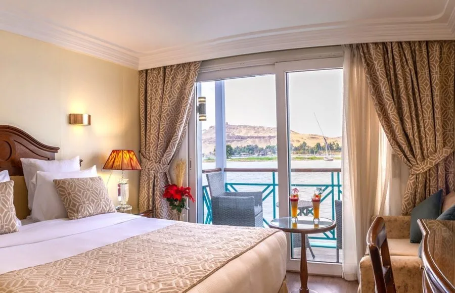 A luxurious suite on the Sonesta Moon Goddess Nile Cruise ship, featuring a comfortable bed, a large window, and a private balcony overlooking the scenic Nile River.