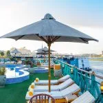 The sunny deck of the Sonesta Moon Goddess Nile Cruise ship, featuring a swimming pool, sun loungers with umbrellas, and a stunning view of the Nile River.