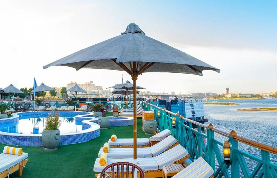 The sunny deck of the Sonesta Moon Goddess Nile Cruise ship, featuring a swimming pool, sun loungers with umbrellas, and a stunning view of the Nile River.