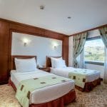 A comfortable twin-bed cabin on the Ms Nile Goddess Nile Cruise with a large window overlooking the Nile River and mountains.