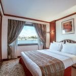 A luxurious suite on the MS Nile Goddess Nile Cruise, with a bed, television, and large window overlooking the Nile River.
