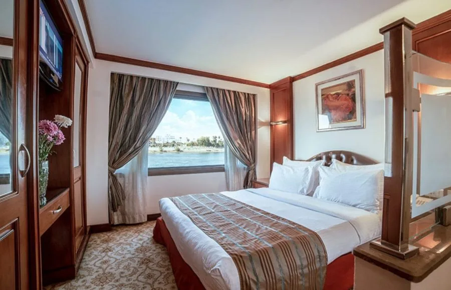 A luxurious suite on the MS Nile Goddess Nile Cruise, with a bed, television, and large window overlooking the Nile River.