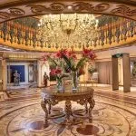 The lavish lobby of the Ms Sonesta St George Nile Cruise, featuring an ornate chandelier, a central marble table with a floral arrangement, and elegant, gilded pillars and staircase.