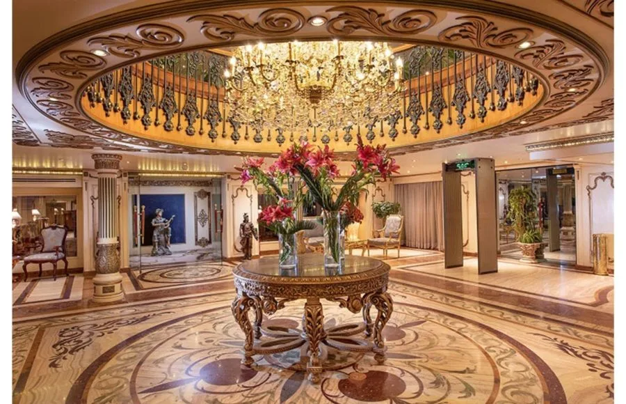 The lavish lobby of the Ms Sonesta St George Nile Cruise, featuring an ornate chandelier, a central marble table with a floral arrangement, and elegant, gilded pillars and staircase.
