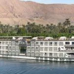 Ms Sonesta St George Nile Cruise ship sailing along the tranquil Nile River with palm trees and a mountain in the background.