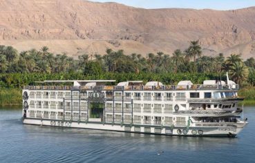 Ms Sonesta St George Nile Cruise ship sailing along the tranquil Nile River with palm trees and a mountain in the background.