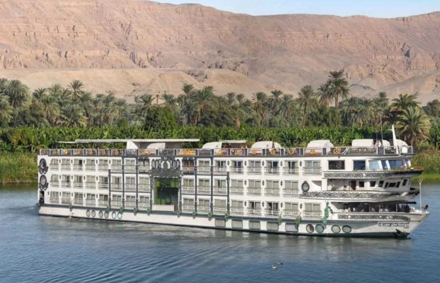 Ms Sonesta St George Nile Cruise ship sailing along the tranquil Nile River with palm trees and a mountain in the background.