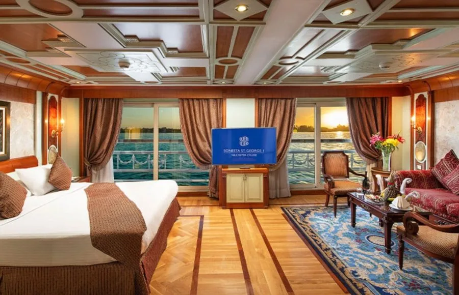 A luxurious suite on the MS Sonesta St George Nile Cruise with a comfortable bed, elegant furnishings, and a large window offering a stunning view of the Nile River.