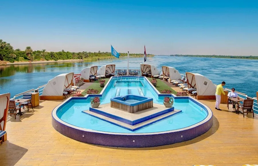 The stunning sun deck of the MS Sonesta St George Nile Cruise ship, featuring a swimming pool, jacuzzi, and comfortable lounge chairs, with the majestic Nile River in the background.