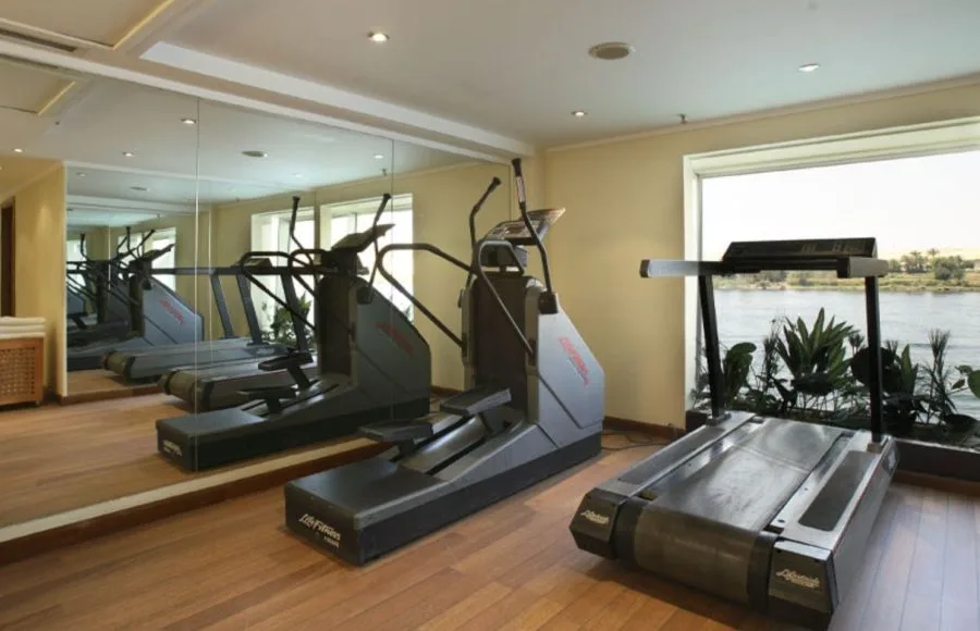 The onboard gymnasium of the Sonesta Star Goddess Nile cruise ship, featuring a treadmill, an elliptical machine, and large windows with a scenic view of the Nile River.