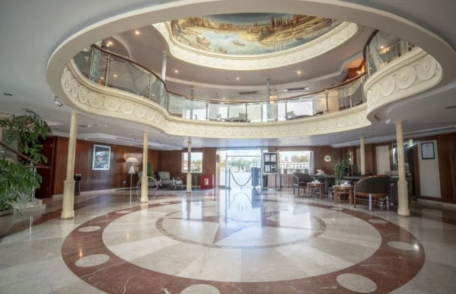 The grand and elegant lobby of the Sonesta Star Goddess Nile cruise ship, featuring a high ceiling with a beautiful mural and a sweeping staircase.