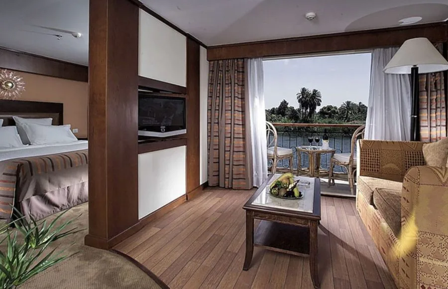 A spacious suite on the Sonesta Star Goddess Nile cruise ship, featuring a comfortable bed, a separate sitting area, and a private balcony overlooking the Nile River.