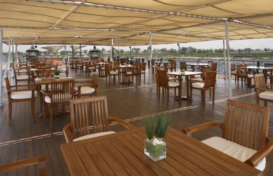 An inviting outdoor dining area on the sundeck of the Sonesta Star Goddess Nile cruise ship, with wooden tables and chairs shaded by a canopy.
