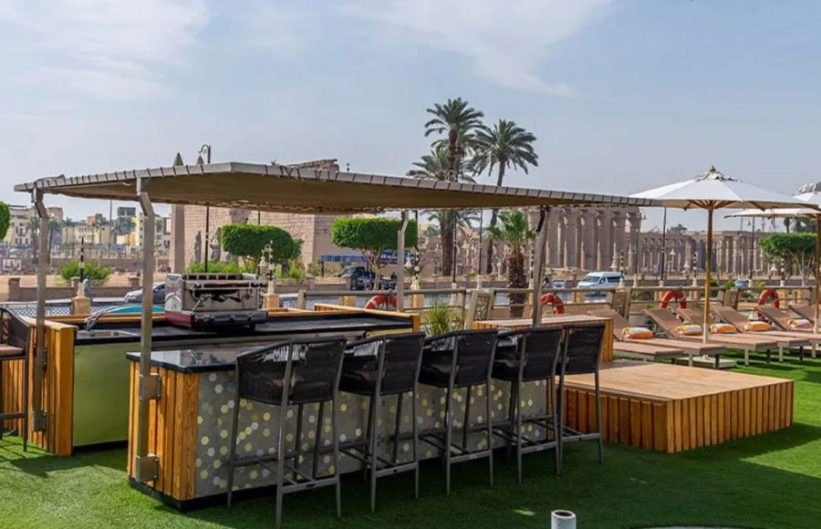 The sunny deck bar on the Sonesta Sun Goddess Nile cruise ship, with comfortable stools and a view of ancient Egyptian ruins in the distance.