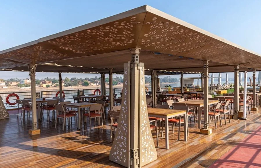 Guests relaxing on the sunny top deck of the Sonesta Sun Goddess Nile cruise ship, enjoying a meal under a patterned canopy with views of the Nile River.