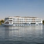 The striking white silhouette of the MS Tamr Henna Nile Cruise glides serenely on the tranquil waters of the Nile with lush greenery and distant desert visible under a clear sky