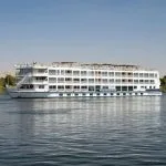 The striking white silhouette of the MS Tamr Henna Nile Cruise glides serenely on the tranquil waters of the Nile with lush greenery and distant desert visible under a clear sky