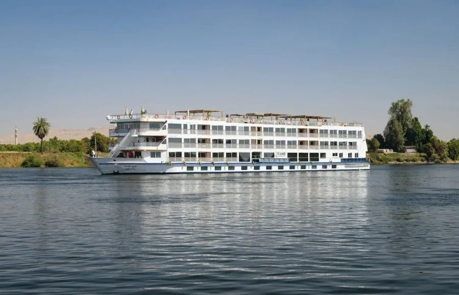 The striking white silhouette of the MS Tamr Henna Nile Cruise glides serenely on the tranquil waters of the Nile with lush greenery and distant desert visible under a clear sky