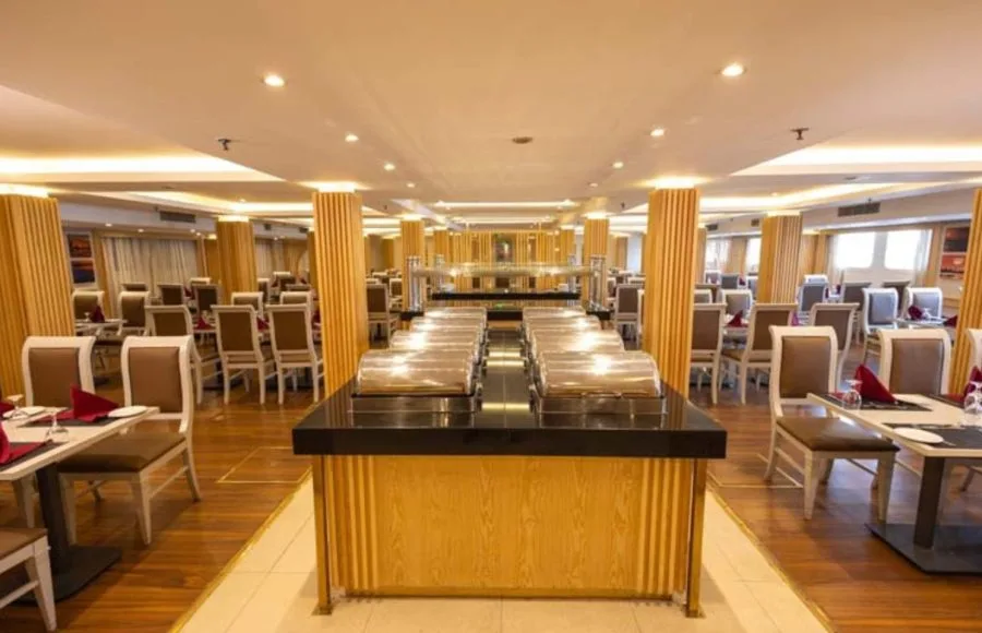 Formal dining room on a ship with round and rectangular tables, high-backed chairs, and wooden columns.