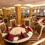 Formal dining room on a ship with round and rectangular tables set with red napkins, surrounded by wooden columns.