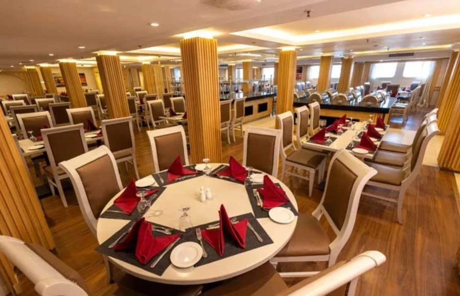 Formal dining room on a ship with round and rectangular tables set with red napkins, surrounded by wooden columns.