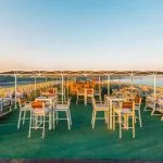 Outdoor bar and lounge area on a ship's top deck at sunset, featuring high tables, white stools, and comfortable wicker-style seating under a partially shaded canopy.