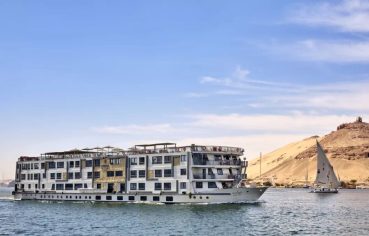 Experience the majestic journey of the MS Tulip Nile Cruise as this elegant ship glides along the historic river with a traditional sailboat and the desert landscape in the background.