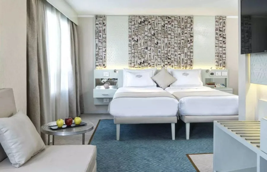 A bright and inviting cabin on the MS Tulip Nile Cruise with twin beds, modern decor featuring hieroglyphics, and a cozy seating area with a fruit platter.