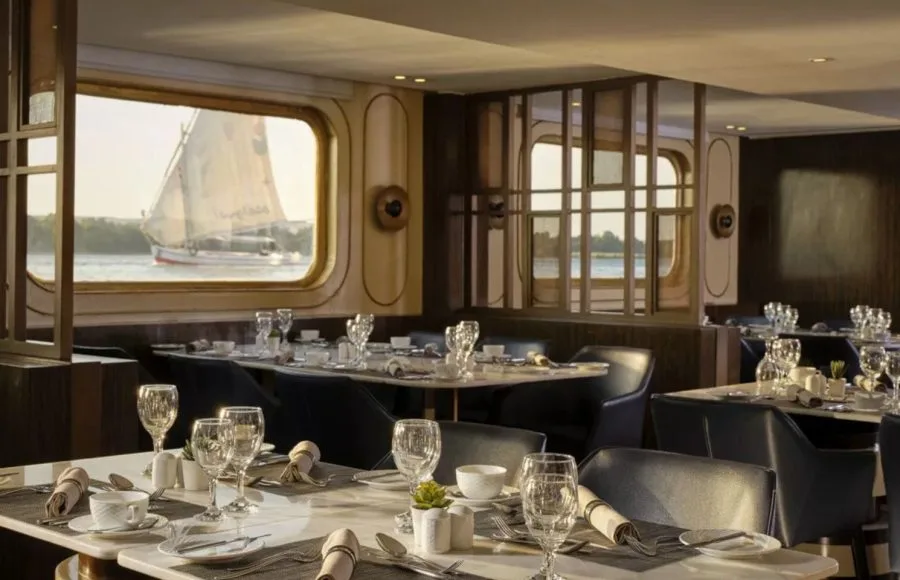 The elegant dining room of the MS Tulip Nile Cruise with set tables and a large window providing an engaging view of a felucca sailing on the Nile River.