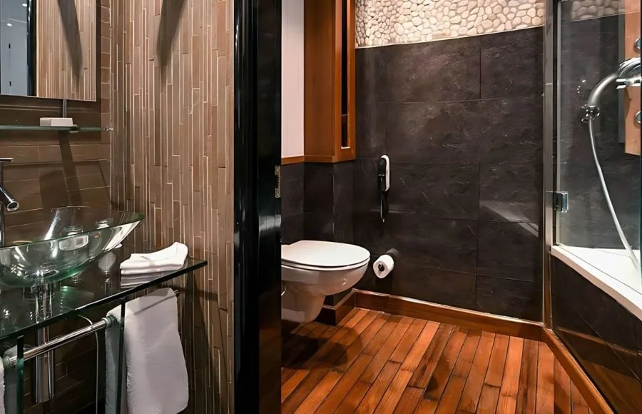 A modern and stylish bathroom onboard the Moondance Nile Cruise, featuring a sleek glass basin, a contemporary toilet, and a spacious shower area with natural wood flooring and elegant stone tiling.