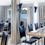 The elegant dining room of the Moondance Nile Cruise, featuring beautifully set tables with black leather chairs and large windows offering scenic views of the Nile River and passing sailboats.