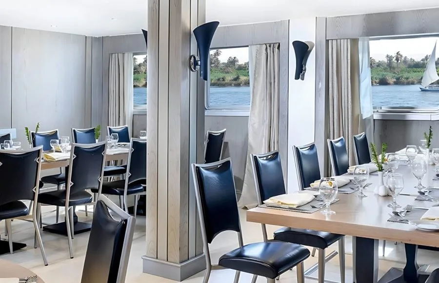 The elegant dining room of the Moondance Nile Cruise, featuring beautifully set tables with black leather chairs and large windows offering scenic views of the Nile River and passing sailboats.