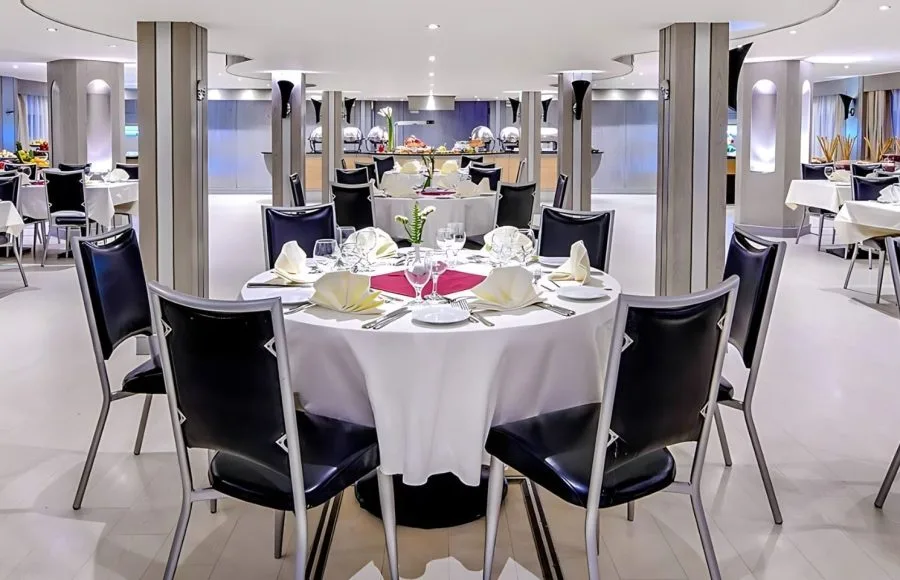 The elegant and spacious restaurant on the Moondance Nile Cruise, featuring a round table with a pristine white tablecloth, black chairs, and a buffet area in the background.