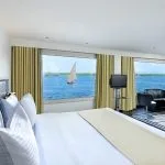 A luxurious cabin on the Moondance Nile Cruise with a king-size bed, a seating area, and large windows offering breathtaking panoramic views of the Nile River and a sailboat.