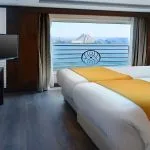 A comfortable cabin on the Sindbad Nile Cruise, featuring twin beds, a television, and a large window offering a stunning view of a felucca sailing the Nile.